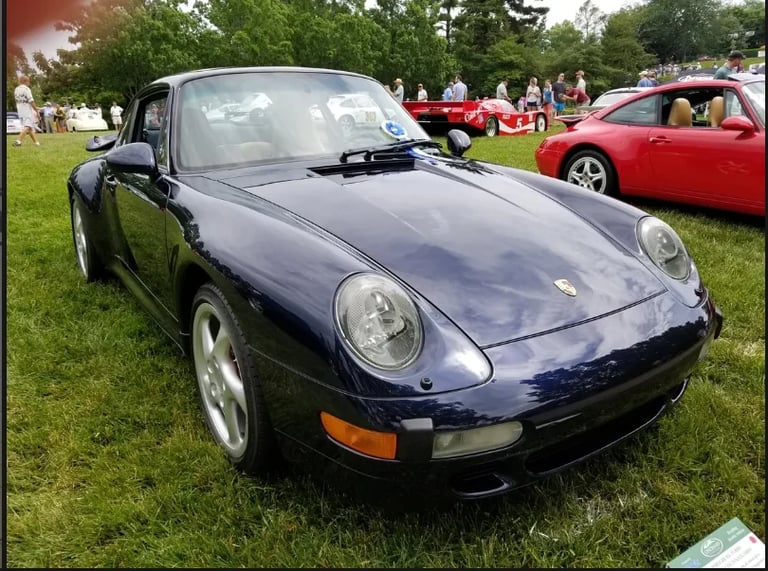 Dark blue Porsche sports car displayed at outdoor car show on grass with other vehicles in background