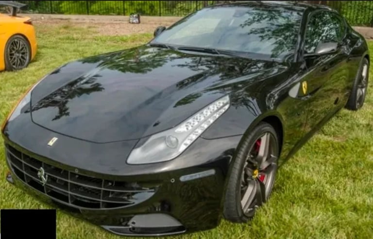Black Ferrari sports car parked on grass at outdoor car show with yellow car visible in background